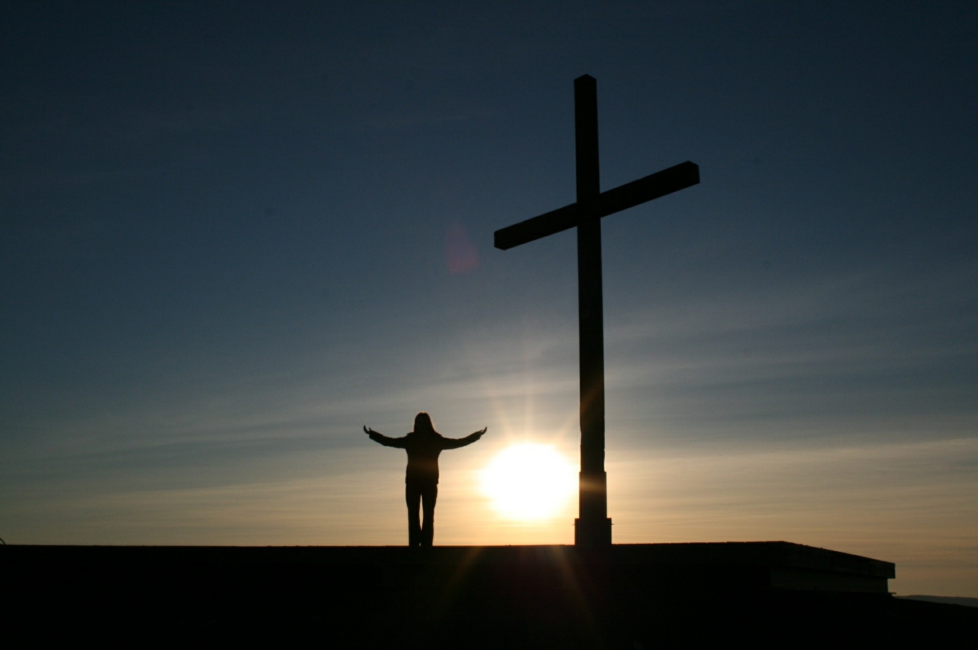 Frau steht neben Gipfelkreuz und wir von vorn von der Sonne angeschienen